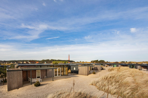 Moderne strandhuisjes tussen duinen en helmgras, met een vuurtoren op de achtergrond onder blauwe lucht.