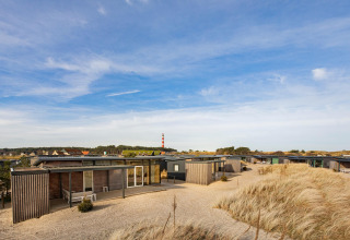 Moderne strandhuisjes tussen duinen en helmgras, met een vuurtoren op de achtergrond onder blauwe lucht.