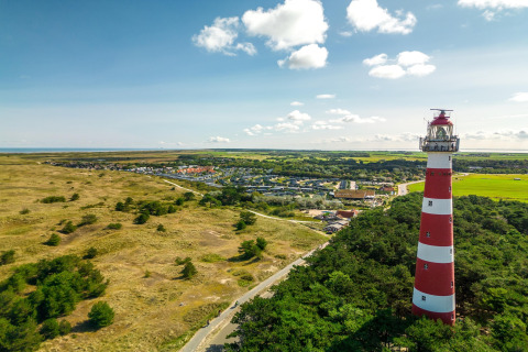 Luchtbeeld van een rood-witte vuurtoren bij een dorp en duinlandschap op een heldere zomerdag.