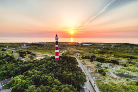 Strandhuis met een rood-witte vuurtoren bij zonsondergang, omgeven door natuur en de zee in de verte.