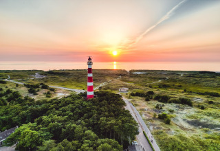 Strandhuis met een rood-witte vuurtoren bij zonsondergang, omgeven door natuur en de zee in de verte.