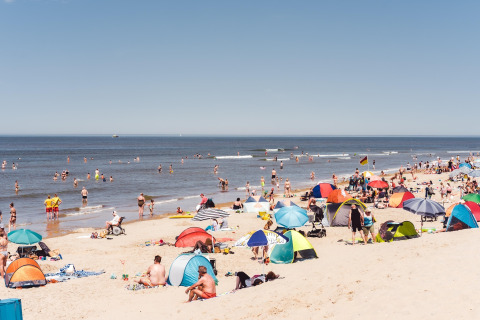 Drukke zomerse stranddag met mensen, kleurrijke parasols en strandtenten langs de zee en blauwe lucht.
