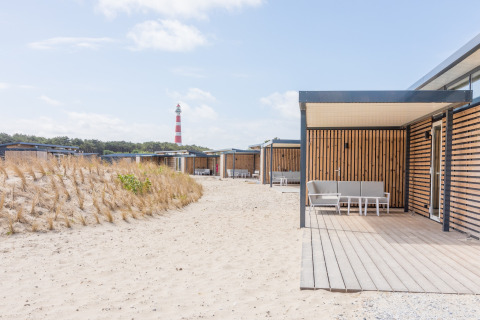 Moderne strandhuisjes met houten panelen en terras, Ameland, Nederland, met vuurtoren op de achtergrond.