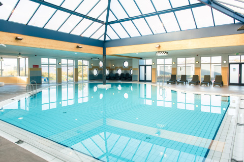 Indendørs swimmingpool med liggestole og store vinduer på et lodge, sollys strømmer ind fra ovenlysvinduer.