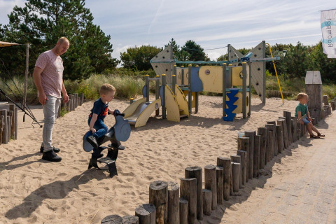 En mand og to børn leger på en legeplads med sand og legeredskaber ved en lodge på en solrig dag.