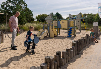 En mand og to børn leger på en legeplads med sand og legeredskaber ved en lodge på en solrig dag.