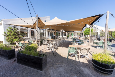Udendørs terrasse på en lodge med borde, stole, planter og solsejl under en blå himmel.