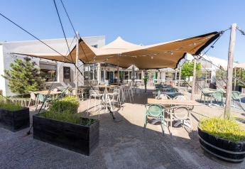 Terraza exterior de un lodge con mesas, sillas, jardineras y toldo bajo un cielo azul despejado.