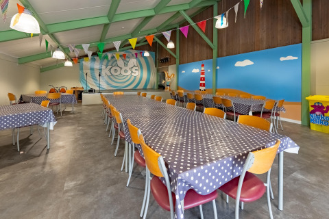 Colorful lodge dining hall with long tables, polka dot tablecloths, bunting, and a beach-themed mural wall.