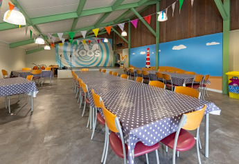Comedor de albergue colorido con mesas largas, manteles de lunares y mural de playa en la pared.