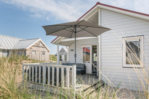 Foto einer gemütlichen Lodge mit Terrasse und Sonnenschirm, helle Fassade und Gräser im Vordergrund.
