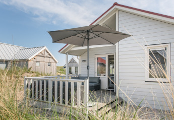Foto af en hyggelig lodge med terrasse og parasol, lys facade og græs i forgrunden på en solrig dag.