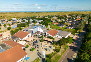 Aerial view of a lodge with red roofs surrounded by holiday cottages and green dunes near the coastline.