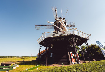 Foto af en traditionel vindmølle ved en lodge på landet under en klar blå himmel, omgivet af marker.