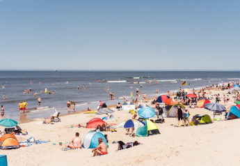 Playa concurrida con muchas personas, tiendas y sombrillas coloridas, clima soleado y mar tranquilo.