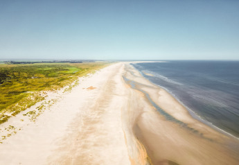 Luftfoto af en lang, uberørt sandstrand ved et lodge med frodige grønne marker og havudsigt.