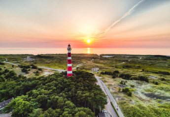 Vista aérea de un faro rojo y blanco junto a un lodge, rodeado de naturaleza y bosque al atardecer.