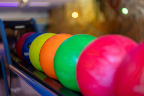 Bolas de boliche de colores alineadas en un soporte en una bolera, con luces de fondo desenfocadas.