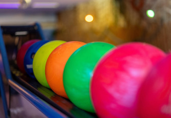 Farverige bowlingkugler står på række i en bowlinghal med slørede lys i baggrunden, fotograferet tæt på.