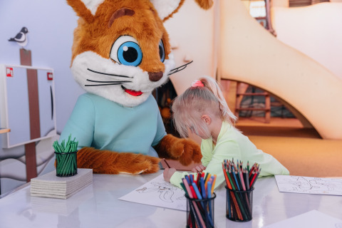 Un bambino colora insieme a una mascotte in costume da animale a un tavolo di un lodge, con matite colorate.