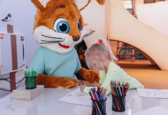 Un niño colorea junto a una mascota disfrazada de animal, sentados en una mesa de una posada con lápices de colores.