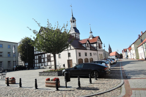 Traditional building and church in Nowe Warpno with parked cars on a cobblestone street on a sunny day.