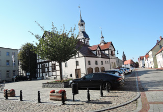 Edificio tradicional y una iglesia en Nowe Warpno con autos estacionados en la calle adoquinada y día soleado.