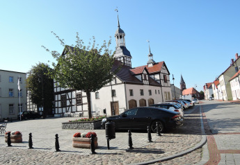 Bâtiment traditionnel et église à Nowe Warpno avec voitures garées sur une rue pavée sous un ciel ensoleillé.