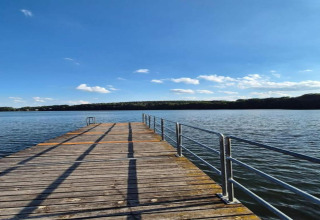 Pont en bois sur le lac devant le Holiday home «Maryna» en Allemagne, sous un ciel bleu et des collines boisées.