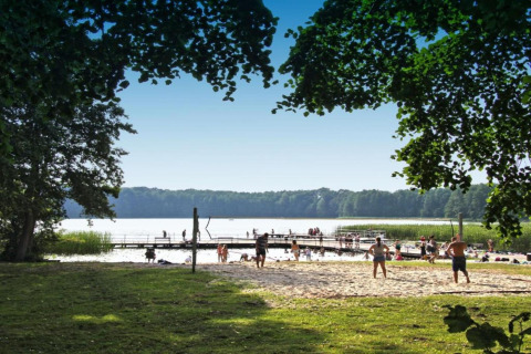 Feriehytte «Maryna» i Tyskland med udsigt over sø, strandvolleyball, badebroer og folk, sommerdag.