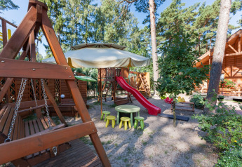 Parque infantil en Domki letniskowe Pobierowo con tobogán, columpios y mesas bajo árboles altos.