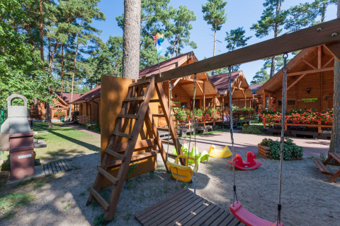 Parco giochi con altalene e casette di legno a Domki letniskowe Pobierowo, tra pini e fiori colorati.