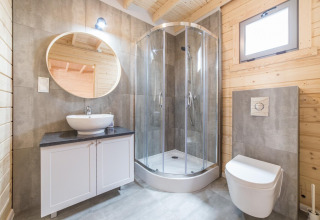 Modernes Badezimmer in einer Holzhütte mit rundem Spiegel, Duschkabine, Waschbecken und WC.