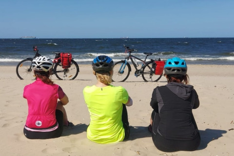 Drie fietsers met helmen zitten op het strand en kijken uit over zee, hun fietsen staan op de achtergrond.