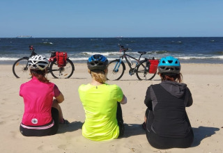 Drie fietsers met een helm zitten op het strand naar zee te kijken, hun fietsen staan op het zand.
