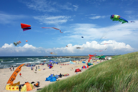 Strand met kleurrijke vliegers en glamping-tenten, blauwe lucht en groene duinen zichtbaar op de voorgrond.