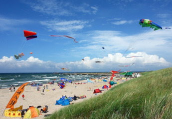 Vista de playa con cometas coloridas y tiendas de glamping, cielo azul brillante y dunas verdes en primer plano.