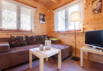 Gezellige houten woonkamer met bruine zetel, kanten gordijnen en tv in Wooden holiday cottages Pobierowo.