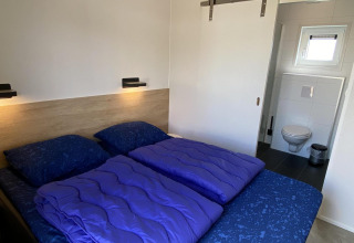 Slaapkamer met tweepersoonsbed, paarse dekens en badkamer in Zonneweelde Lodge Deluxe, Nederland.