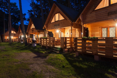 Avondbeeld van verlichte houten vakantiehuisjes met veranda’s bij «Wooden holiday cottages Pobierowo» in Duitsland.