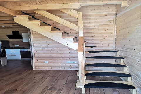 Escalera de madera con peldaños negros en una cabaña de madera, cocina y paredes visibles al fondo.