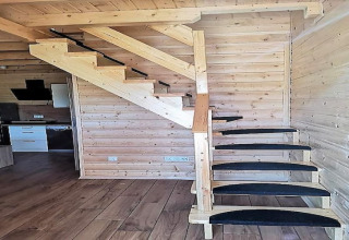 Escalera de madera con peldaños negros en una cabaña de madera, cocina y paredes visibles al fondo.