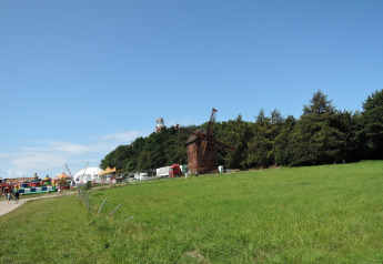 Groene weide met een windmolen, bomen en een kleurrijke kermiszone onder een stralend blauwe hemel.