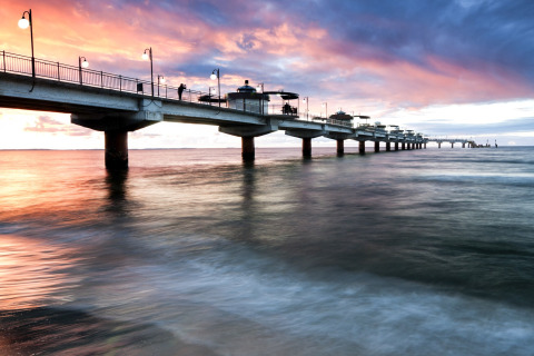 Afbeelding van een lange verlichte pier bij zonsondergang met mensen erop, uitgestrekt over de kalme zee.