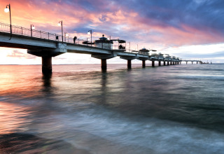 Afbeelding van een lange verlichte pier bij zonsondergang met mensen erop, uitgestrekt over de kalme zee.