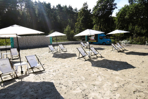 Ligstoelen en parasols op zand bij Turkusowa osada Wapnica, met een blauwe foodtruck nabij het bos.