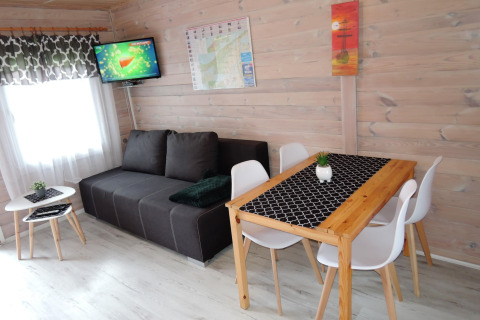 Living area in a German cabin with black sofa, dining table for four, white chairs, and wall-mounted TV.