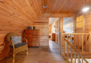 Wooden lodge interior with stairs, wicker chair, dresser, books, and a bedroom visible in the background.