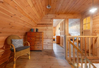 Interno di un lodge in legno con scala, sedia in vimini, cassettiera, libri e camera visibile sullo sfondo.
