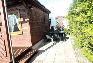 Buitenpatio bij een houten chalet met tafel, stoelen en parasol, omringd door groen onder de zon.